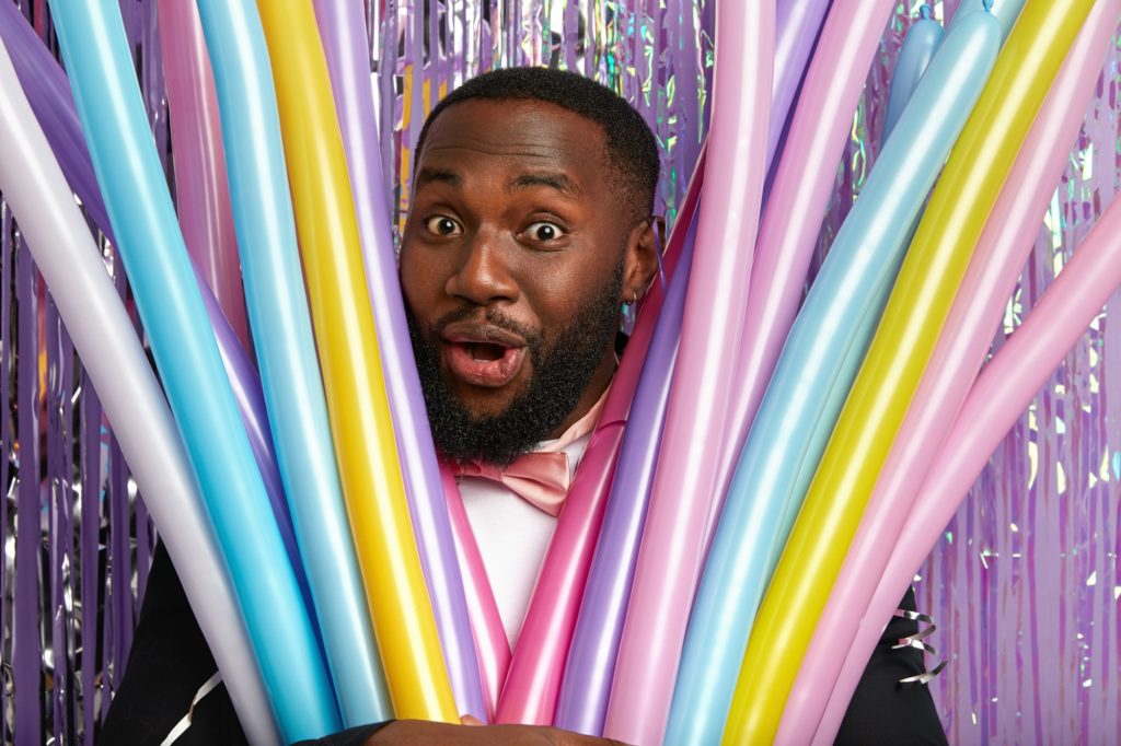 Close up shot of surprised cheerful Afro American guy looks through colorful modeling balloons, has